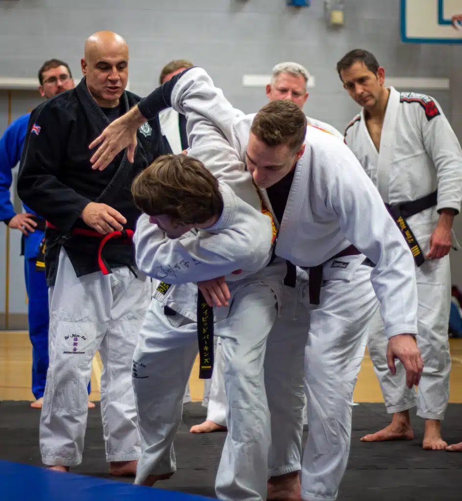 Instructor guiding two students through a jujutsu throw while black belts look on