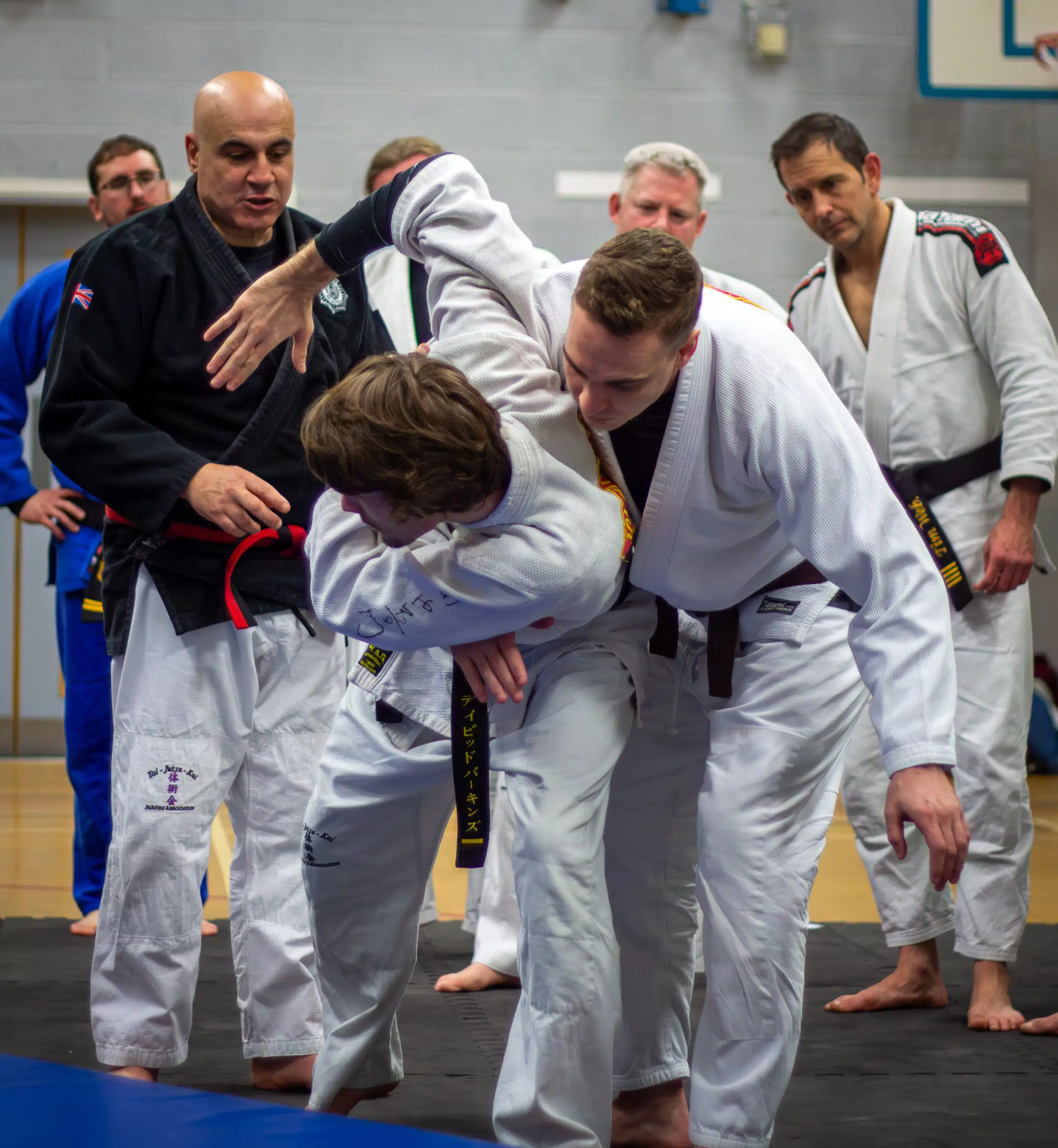 Instructor guiding two students through a jujutsu throw while black belts look on
