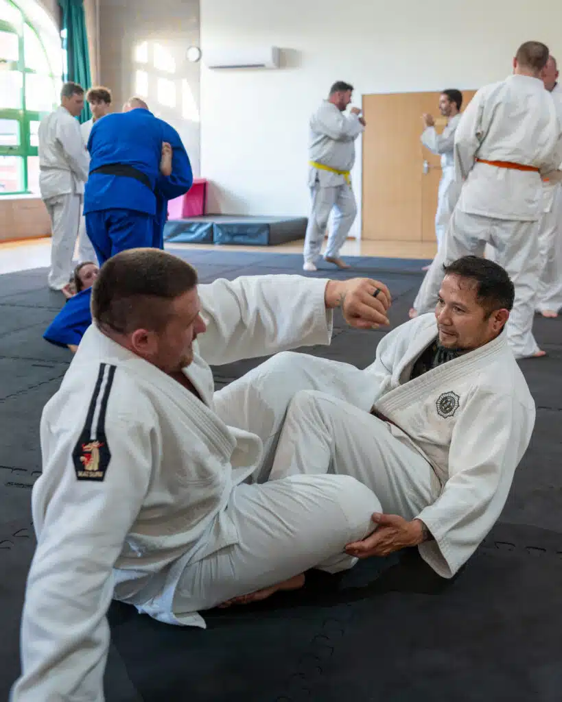 Two students in gis grappling from seated guard while others train behind