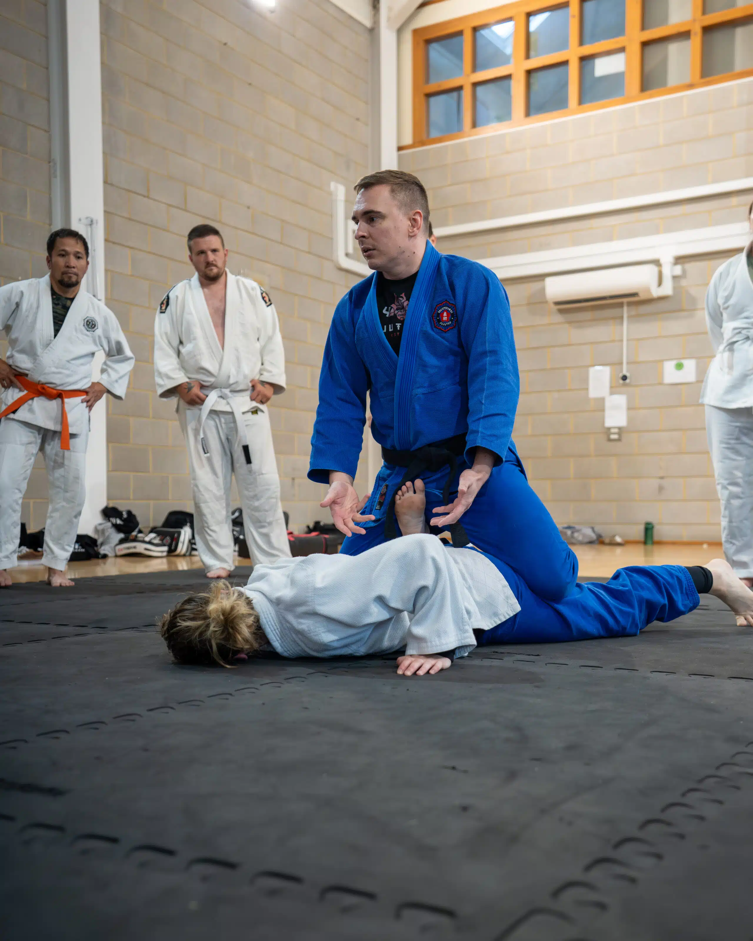 Sensei Mark in a blue gi pinning a student on the mat while the class watches